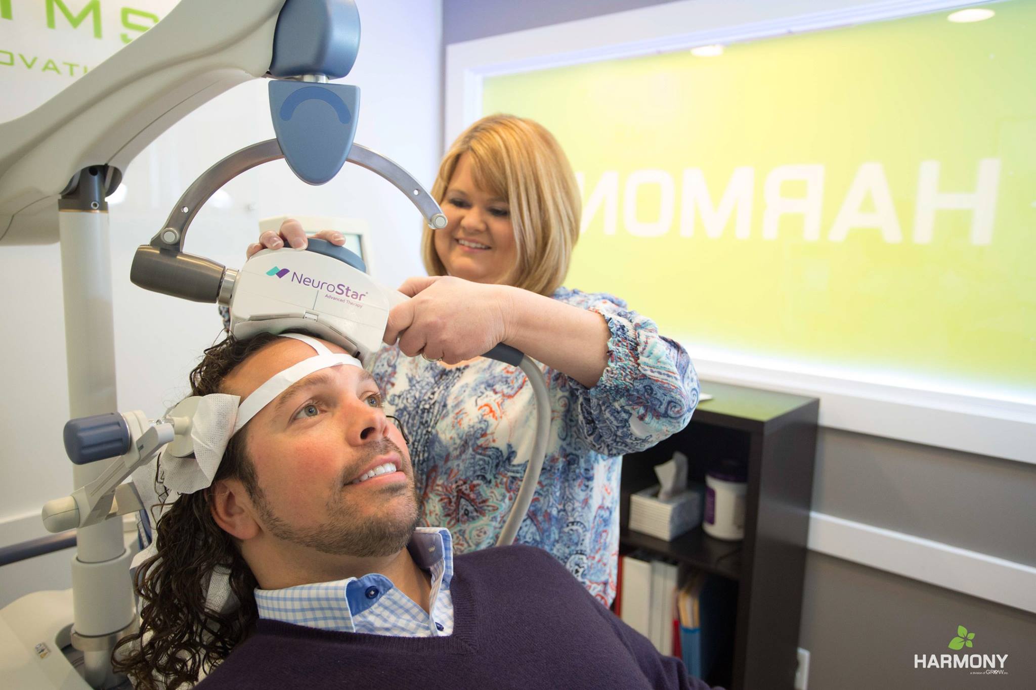 A man in a purple sweater reclines in a chair as a woman with short blonde hair, the tms technician, places the neurostar tms want against his head.
