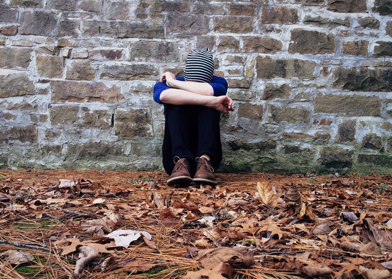 A teen sits against a stone wall on ground covered in fallen leaves. He is wearing a blue sweater with the sleeves pushed up to his elbows and a black and white striped beanie. His legs are pulled to his chest, and his head rests on his knees.