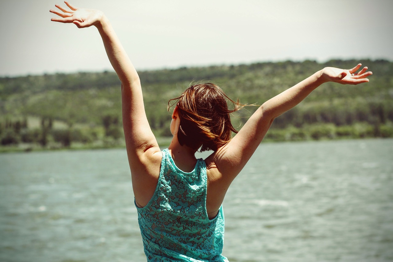 A woman with short brown hair wearing a blue tank top looks out over a lake, her arms outstretched overhead in celebration.