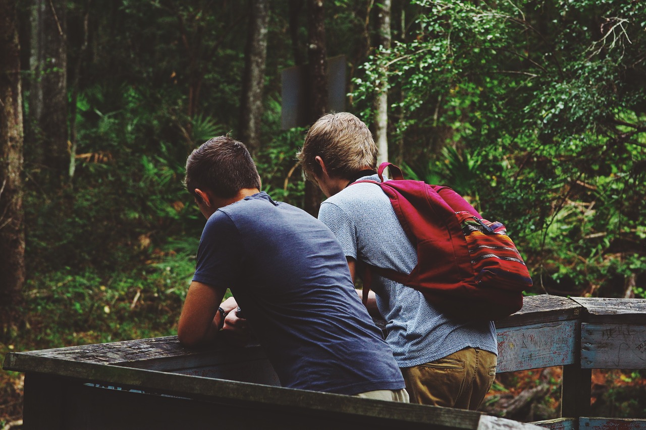 Two young men pause a hike to lean against a railing in a forested area.