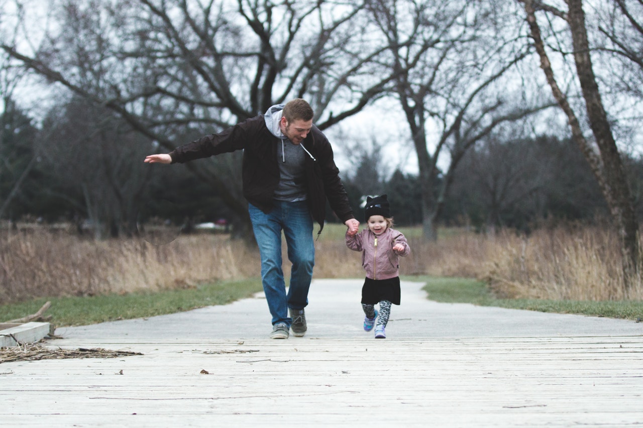 A man and his daughter, a toddler, run together down a path holding hands