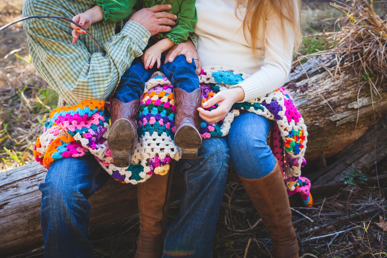 A man and woman sit on a fallen tree branch, a colorful knit blanket spread across their legs, holding their toddler between them.