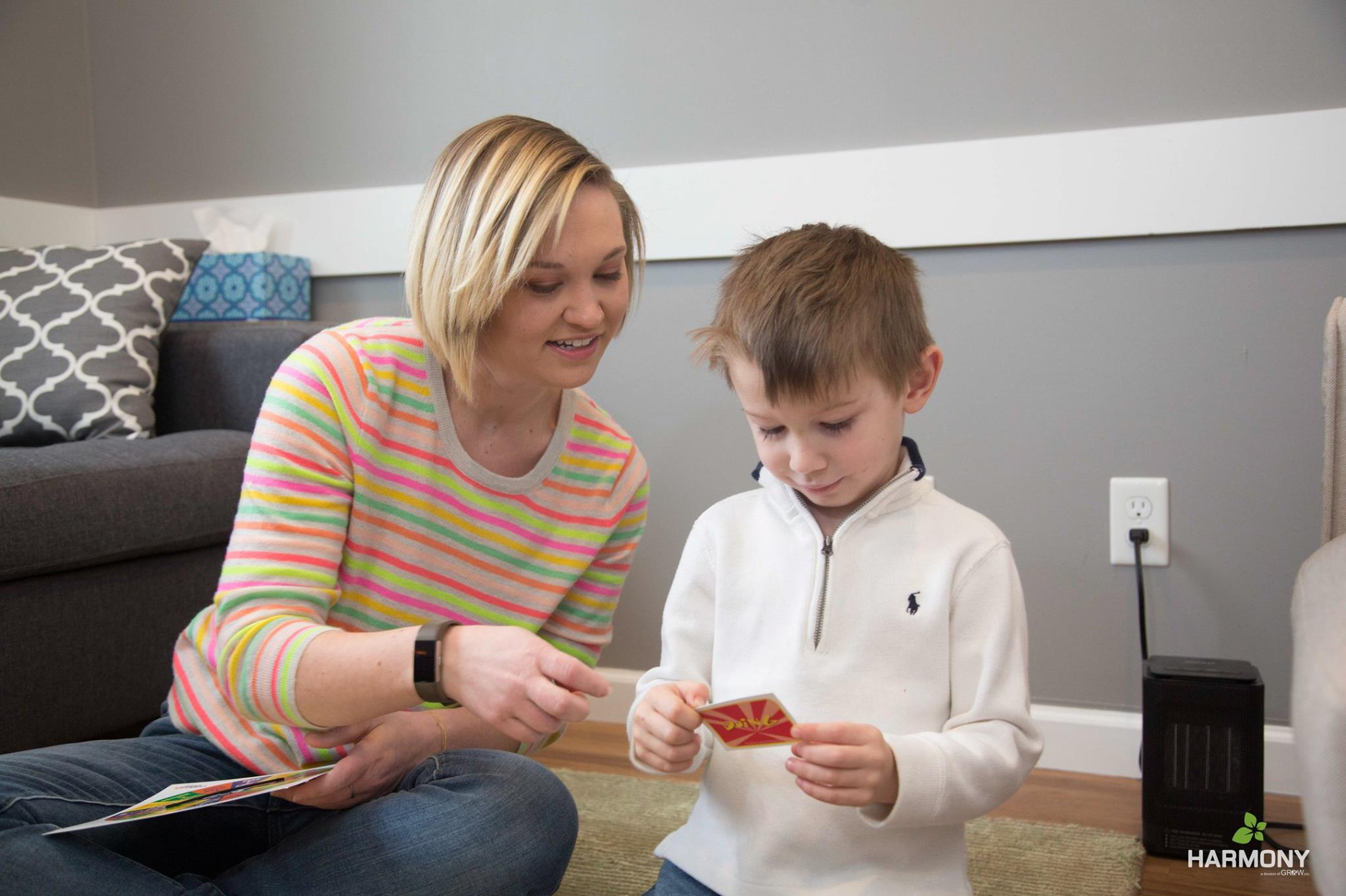 A Harmony counselor with short blonde hair wearing a beige sweater with color stripes looks at a card with a young boy in a white quarter zip.