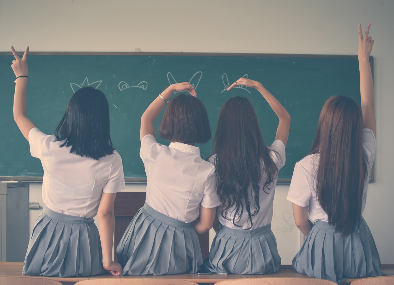 Four girls with brown hair wearing school uniforms lean against a desk with their backs to the camera, facing a chalkboard.