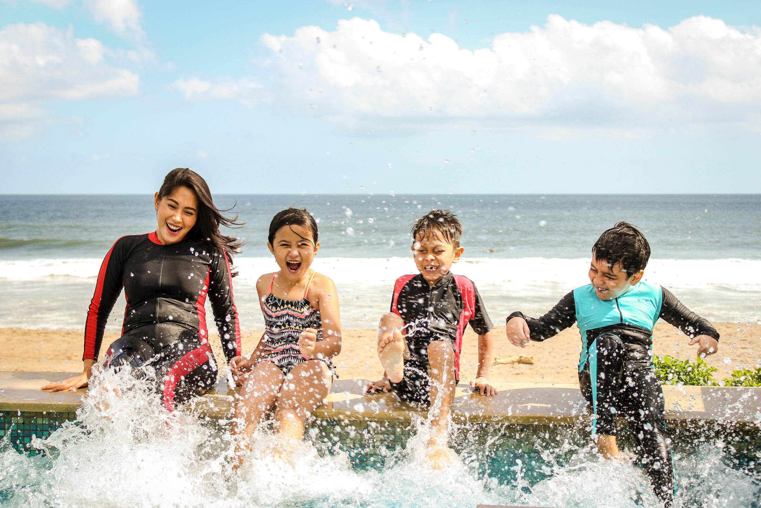 A woman and there children sit at the edge of a beachfront pool, splashing their legs in the pool water.