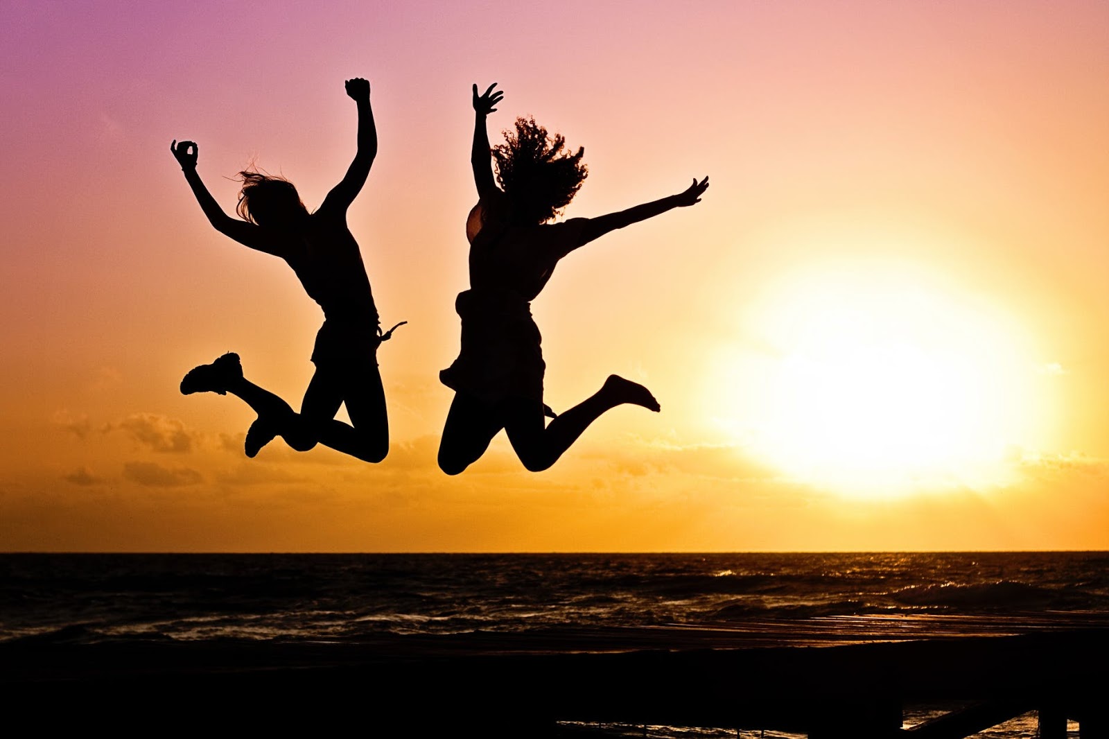 Two people are jumping in the air on a beach as the sun sets behind them. 