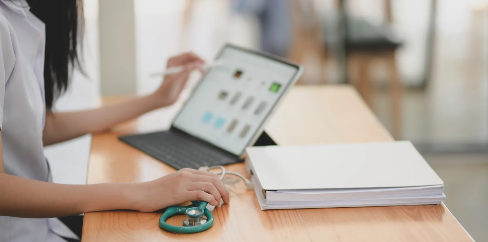 A female medical practitioner, a stethoscope on the table beside her, uses a tablet to work with her patients