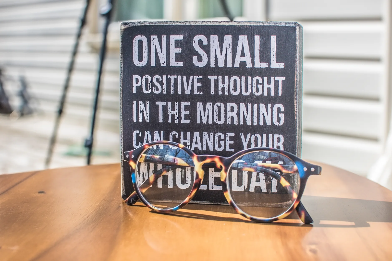 A sign standing on a table has the words "One Small Positive Thought in the Morning Can Change Your Whole Day." A pair of glasses rests in front of it