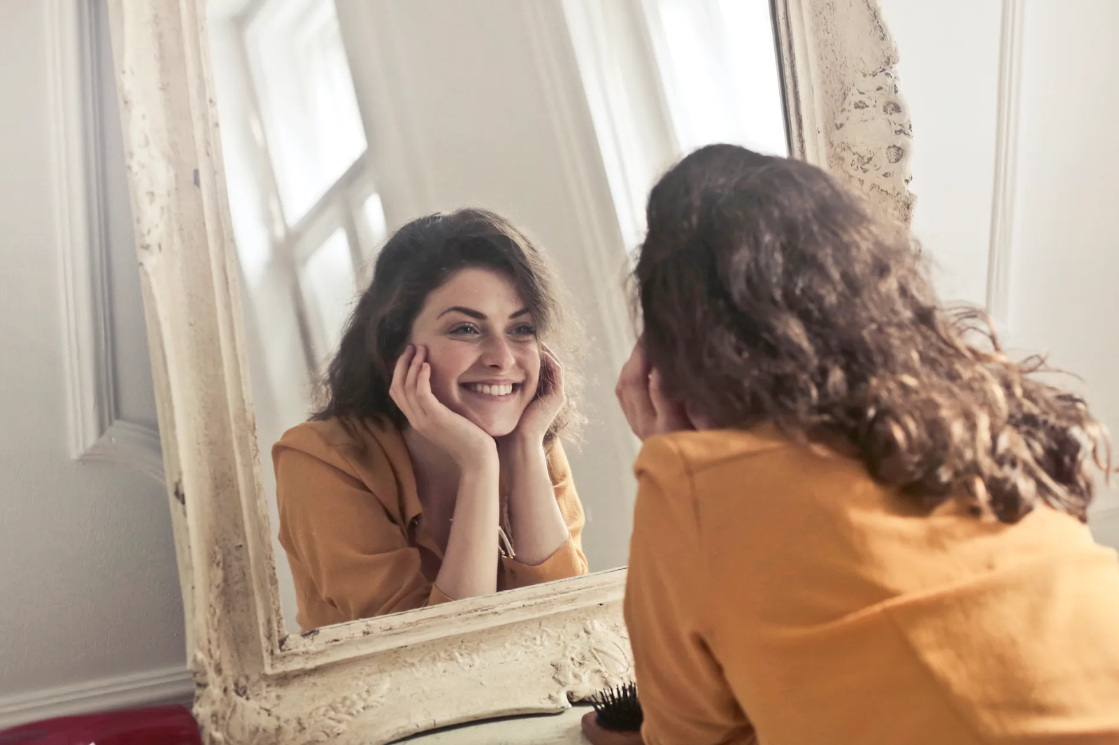 A woman wearing a yellow shirt cups her face as she smiles at her reflection in an ornate white mirror 