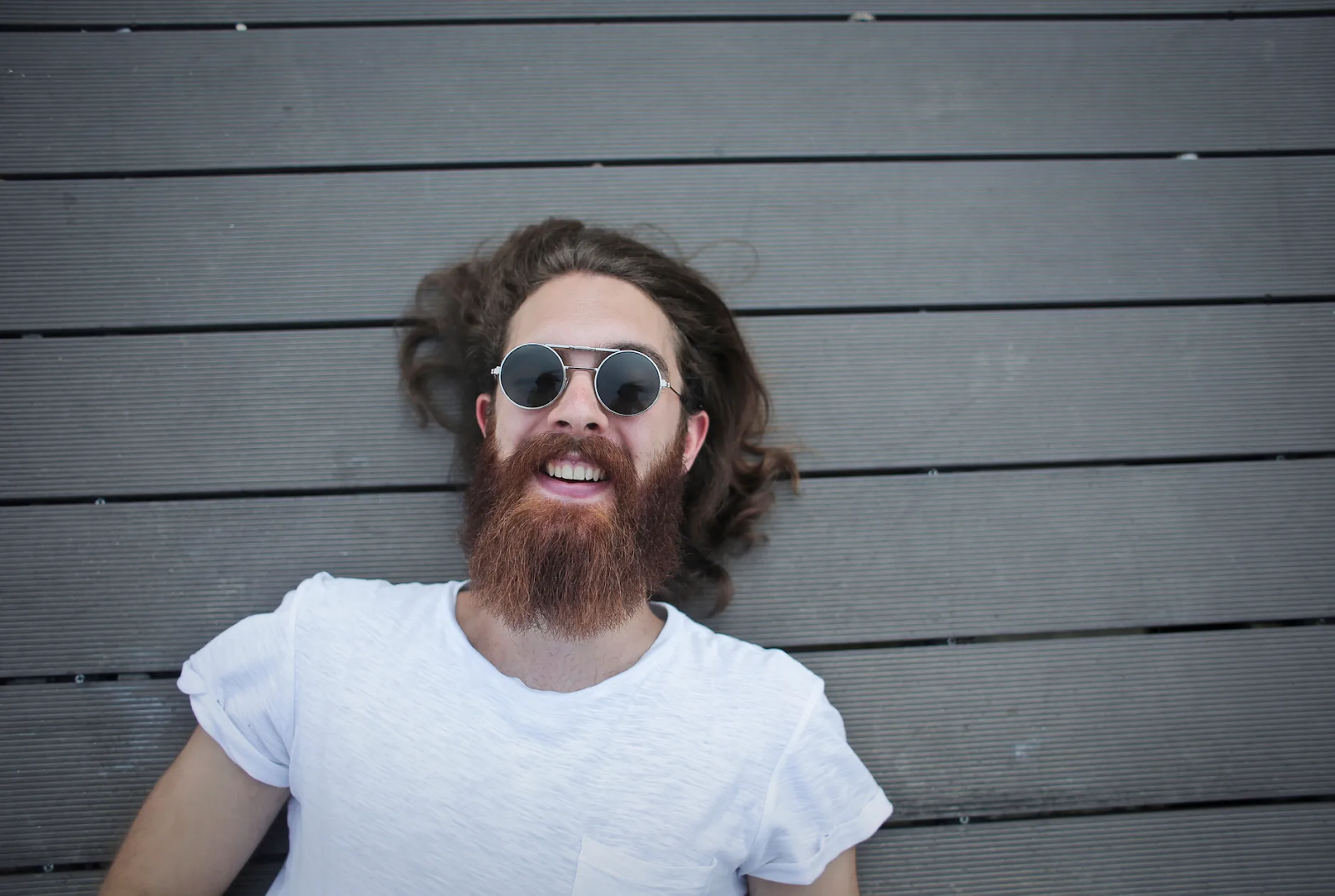 A bearded man wearing sunglasses laughs up at the sky as he lies across gray wooden planks