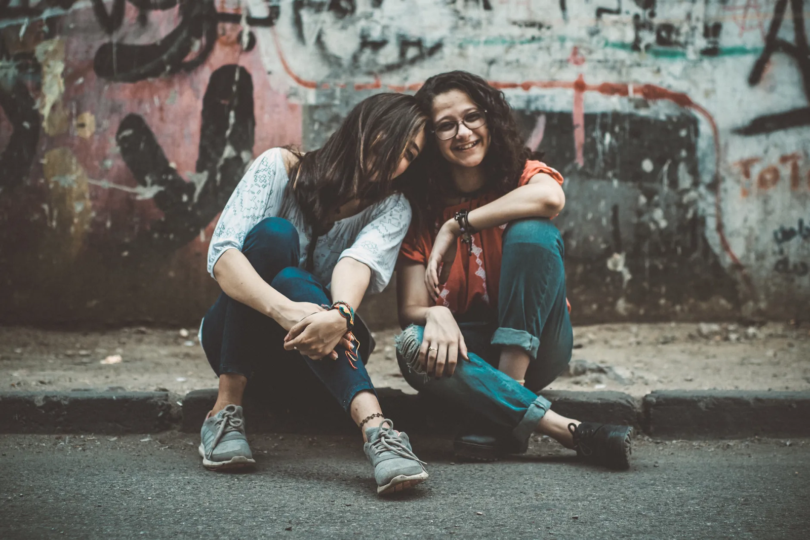 Two woman sit on a curb together in front of a graffiti-covered wall, leaning on each other and laughing