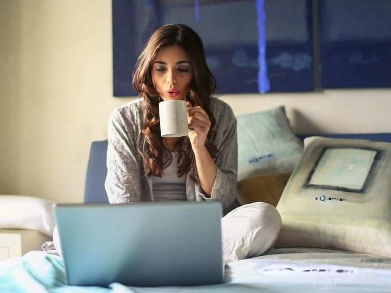 A woman drinks coffee in front of her laptop in bed.