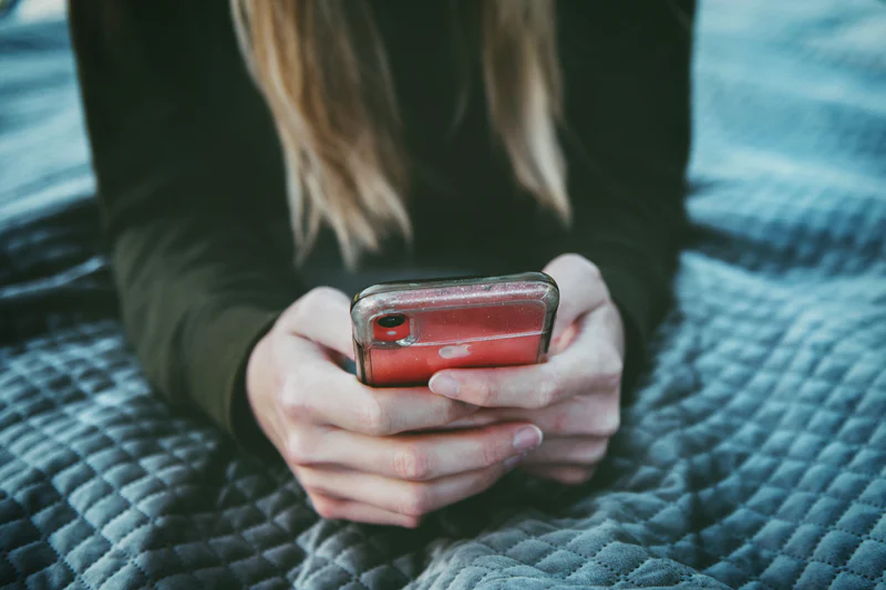An image of a woman under a blanket holding a smartphone in her hands, her face not visible.