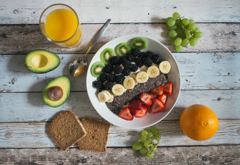 An image depicting a bowl of fruit and granola on a table, along with orange juice, an avocado, bread, and grapes.