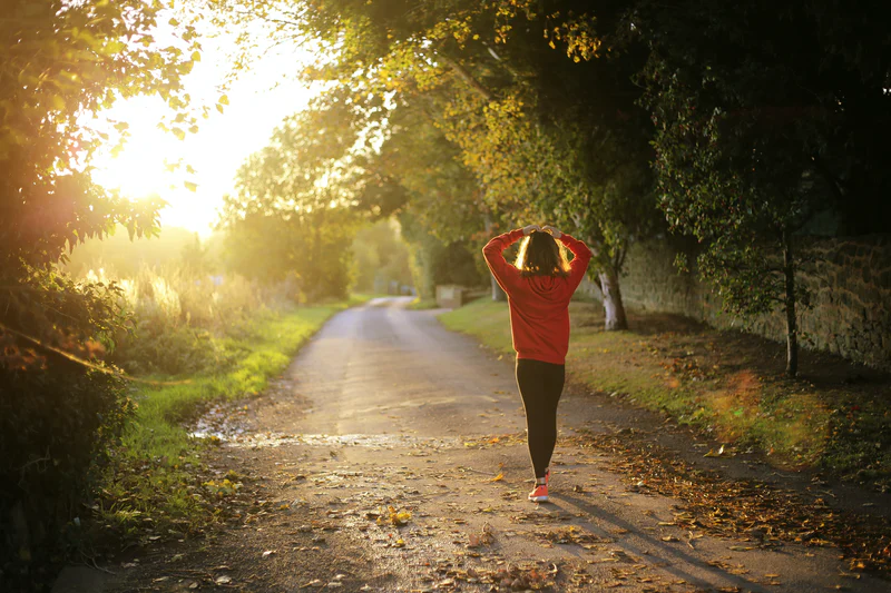 An image of a person walking down a dirt path framed with trees on a sunny day, their hands on their head.