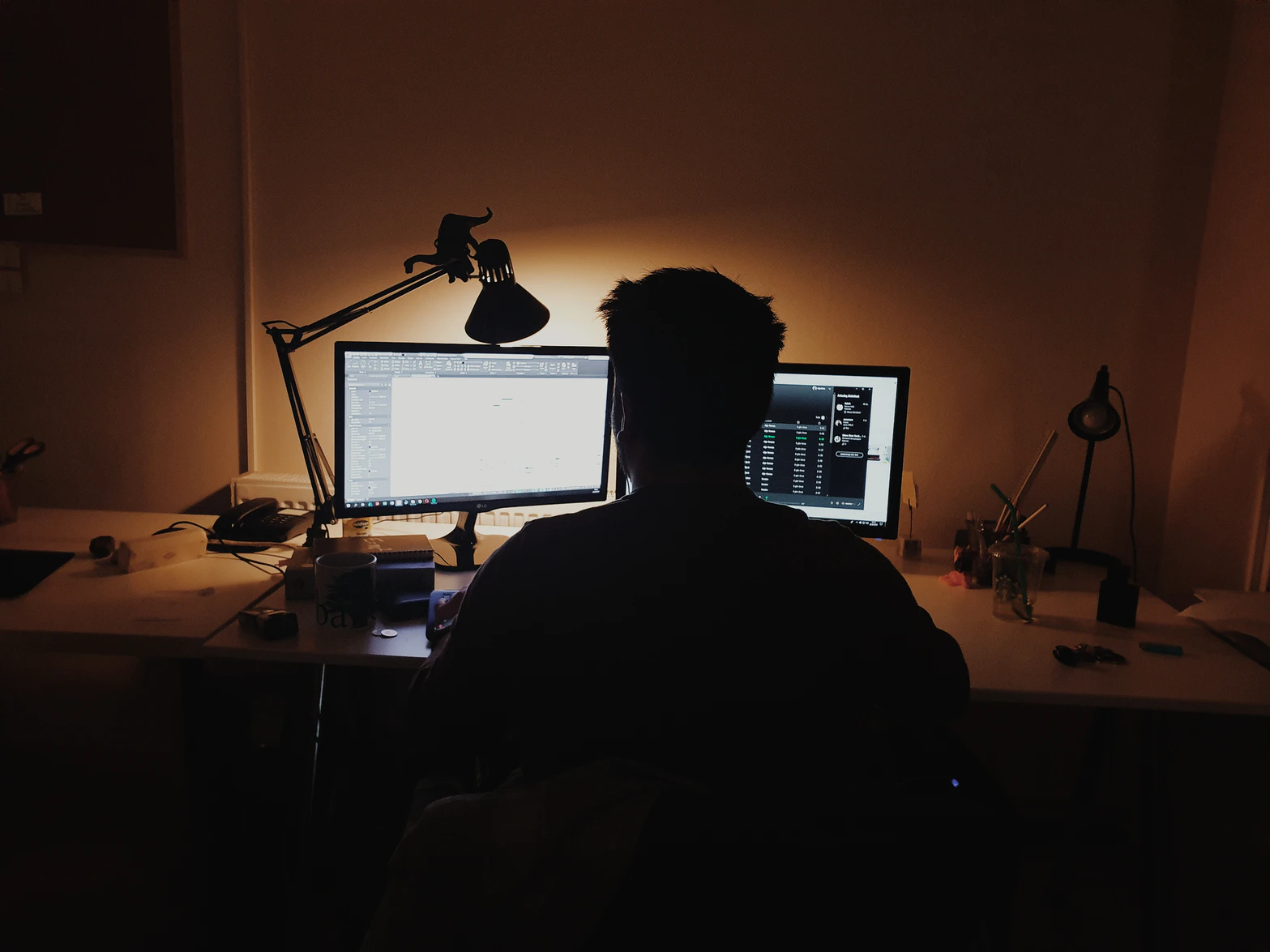 An image of a man sitting alone in a dark room, facing two computer screens.