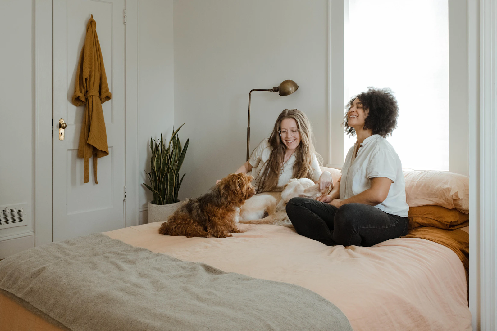 An image of two women sitting on a bed and laughing. One of them is petting a dog that sits beside them.