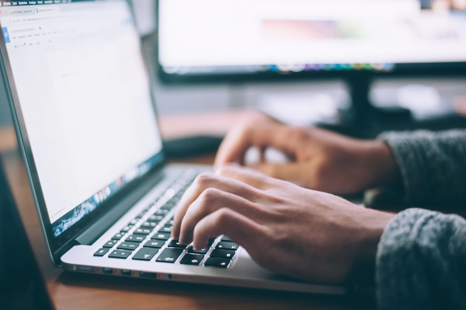 A close-up image of someone's hands typing on a laptop, the screen some sort of document that isn't in focus. Another computer monitor is in the background.