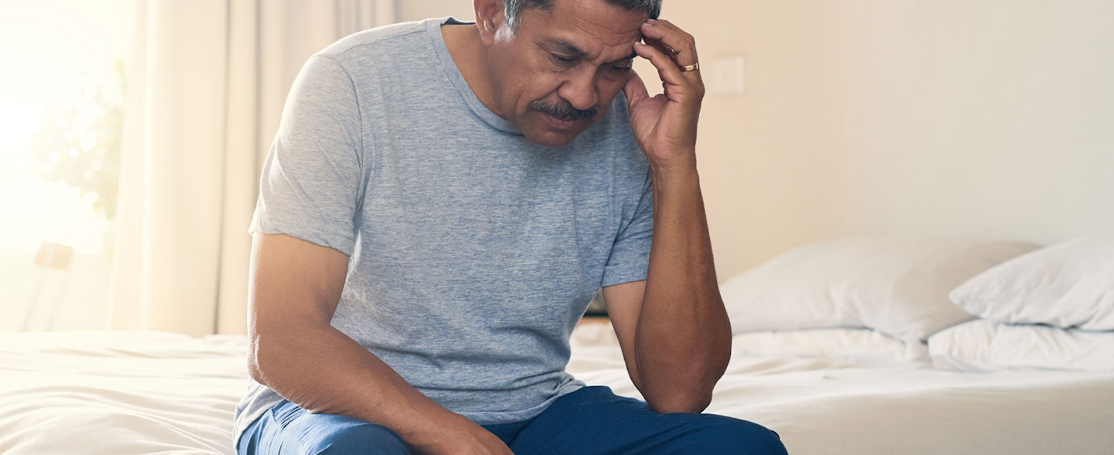 A middle aged man feeling confused from COVID-19 brain fog sits on the edge of his bed.