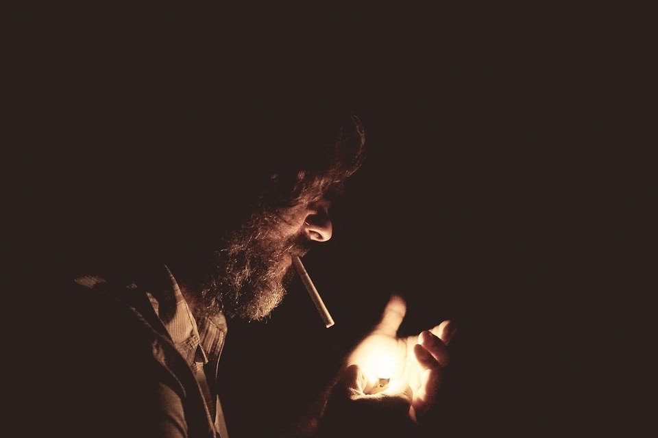 An image of the profile of a bearded man standing in the dark, holding a lighter in his hand to light the cigarette he's holding in his mouth.