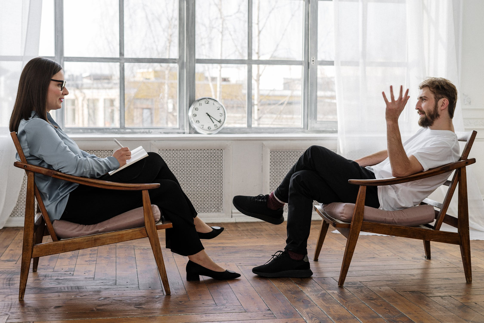 An image of two people in chairs sitting across from each other, with a clock balanced on the window between them. The woman is taking notes and listening as the man talks and gestures.