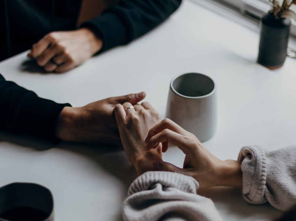 An image of two people holding hands across a table. A cup of coffee sits beside their clasped hands, and a plant is on the edge of the frame.