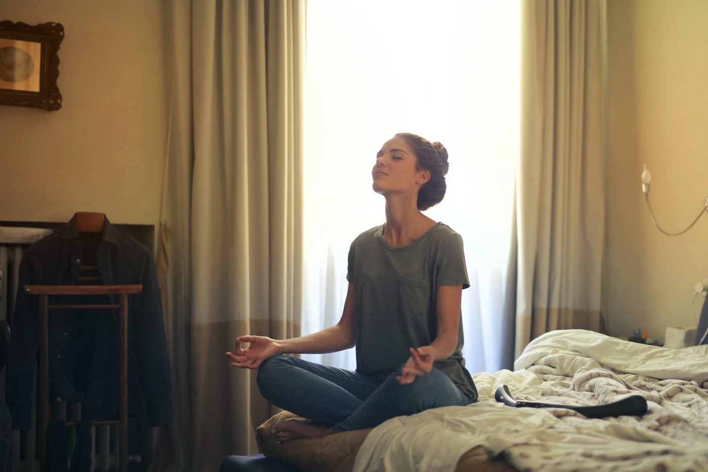 A woman sitting on the edge of her bed meditating while sitting cross-legged.